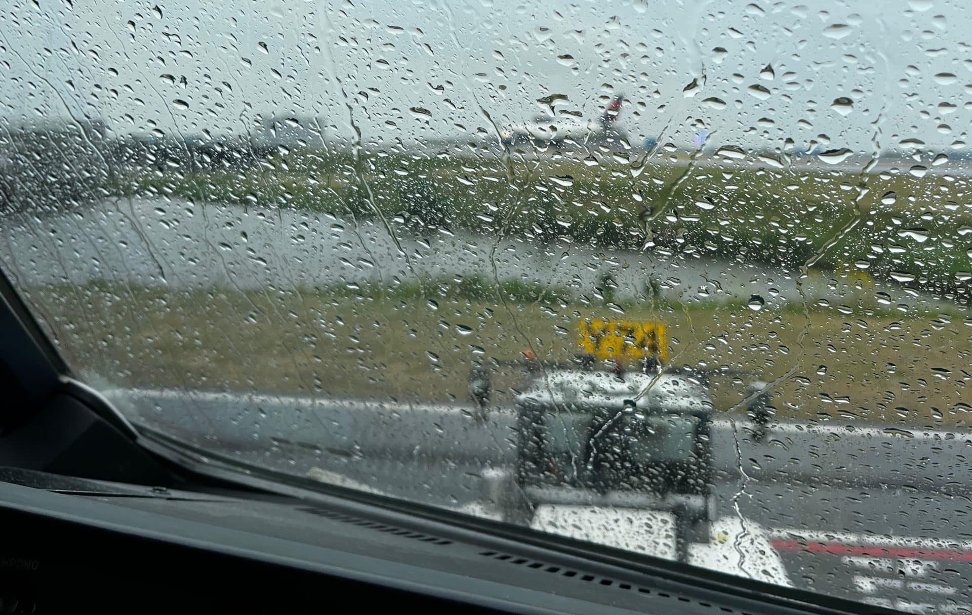 View from aircraft cockpit during cockpit preparation on a rainy day on airport apron, showing raindrops on windshield and a blurred airport runway under overcast skies, highlighting challenging weather conditions.